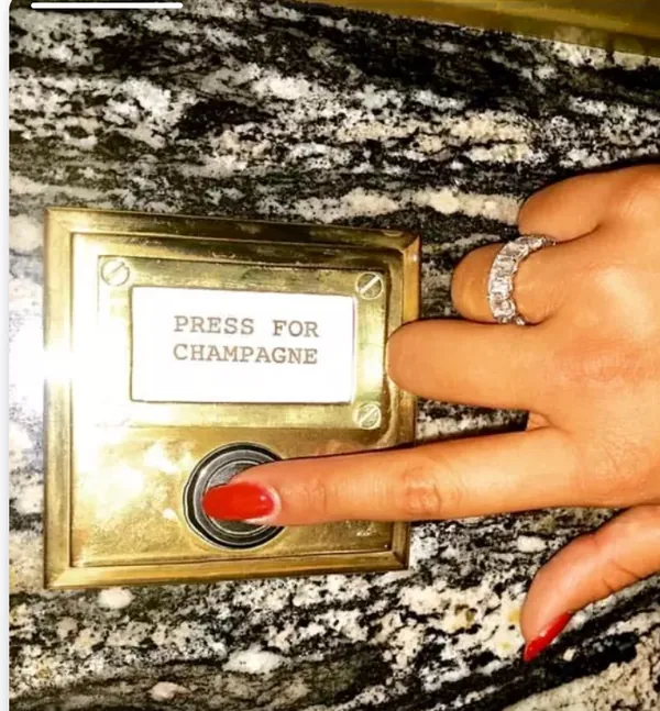 A brass plate in a brick wall engraved PRESS FOR CHAMPAGNE, with a red button beneath it and a hand reaching for it