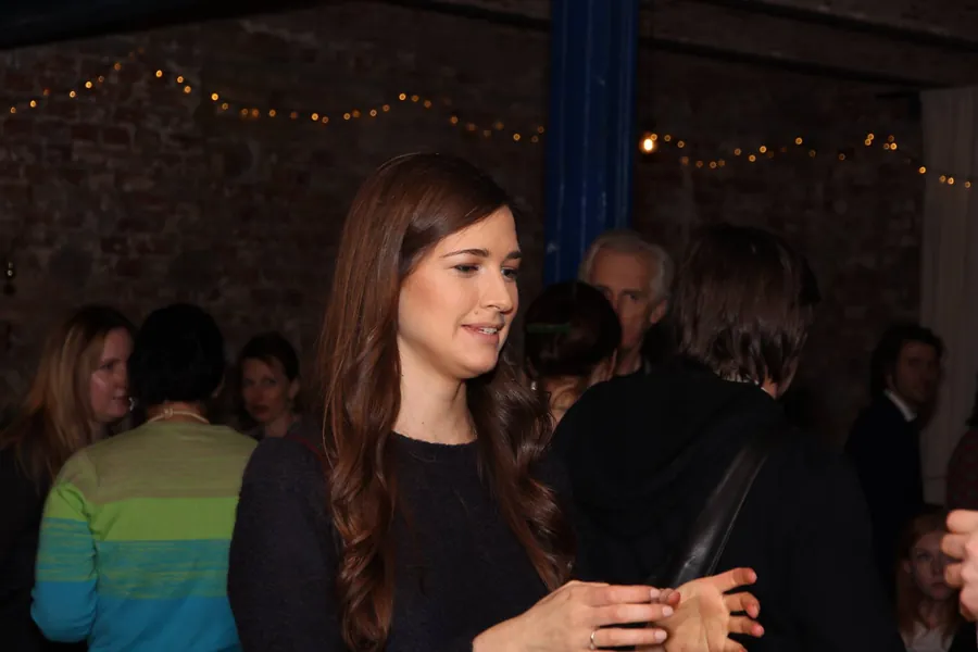A woman with long dark hair talking, glass in hand, the room full behind her