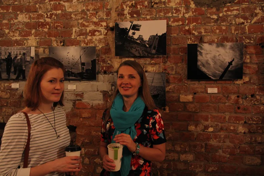 Two women with paper cups by the brick wall, one in a turquoise scarf